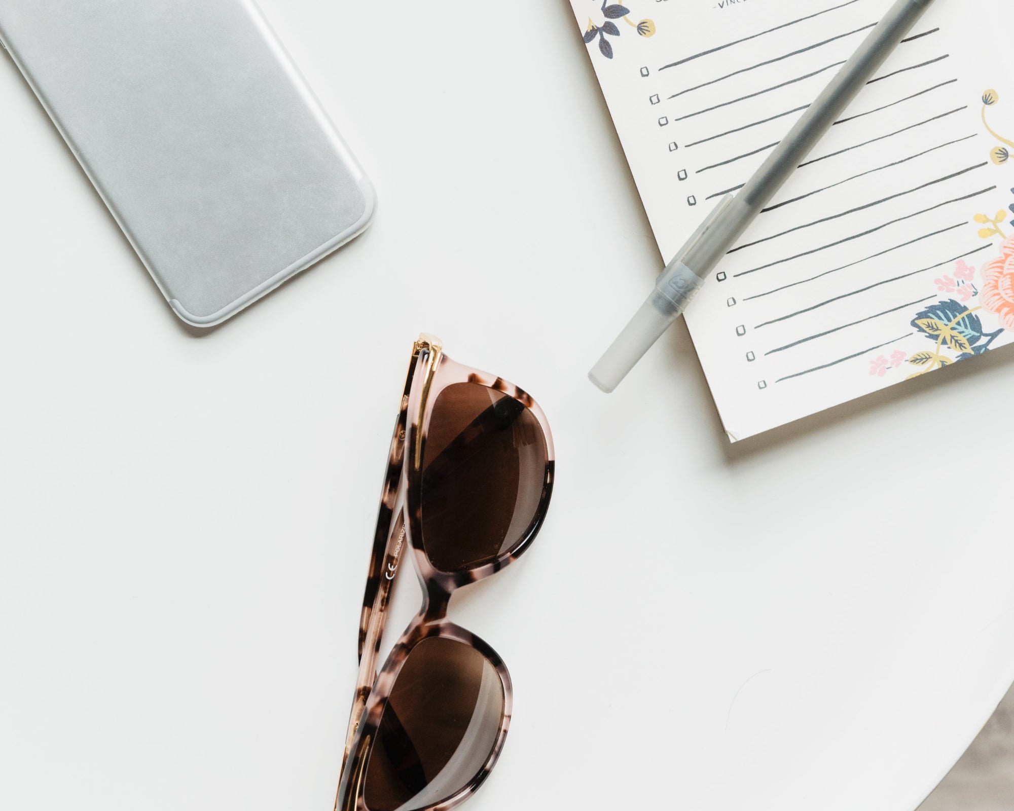 Phone, sunglasses, and planner with pen on a white surface